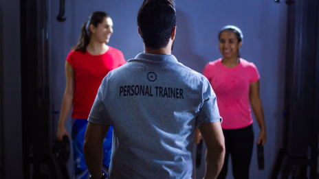 Back view of a personal trainer guiding two women during a gym session indoors.